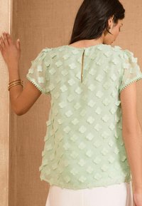 Blusa verde chiaro realizzata in tessuto trasparente, con applicazioni floreali testurizzate, maniche corte e chiusura a oblò sul retro con bottone.