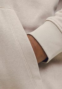 Lys beige sweatshirt med en struktureret overflade, der har ribbede manchetter og en sidelomme med synlige syninger.