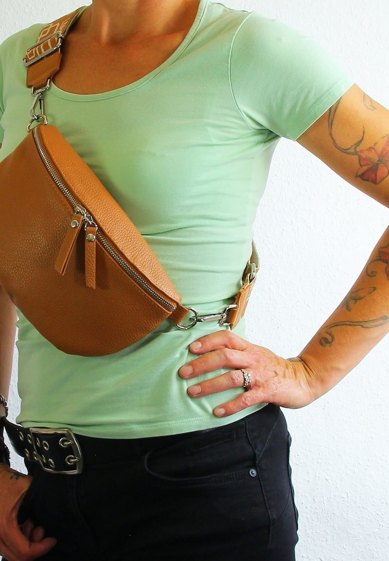 Bruine leren schoudertas met een gestructureerde afwerking, voorzien van een ritssluiting en een verstelbare strap, gedragen over een lichtgroen shirt.