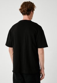 Sort oversized T-shirt med korte ærmer, rund hals og slidseri i kanten. Lavet af bomuld med en glat tekstur. Billede set fra bagsiden.