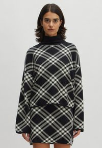 Übergröße schwarzer Strickpullover mit einem weißen und grauen Karomuster, mit hohem Kragen und weiten Ärmeln, kombiniert mit einem passenden Rock.