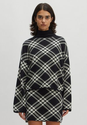 Übergröße schwarzer Strickpullover mit einem weißen und grauen Karomuster, mit hohem Kragen und weiten Ärmeln, kombiniert mit einem passenden Rock.