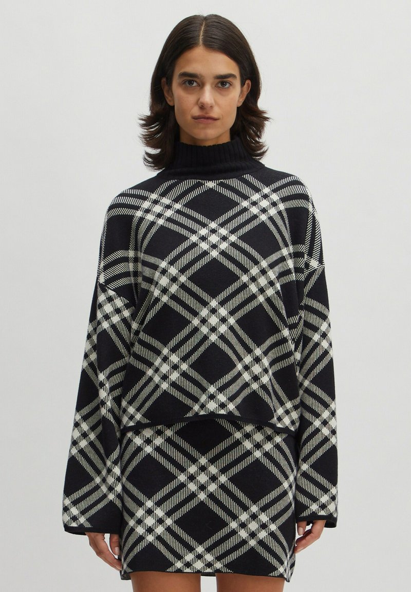 Übergröße schwarzer Strickpullover mit einem weißen und grauen Karomuster, mit hohem Kragen und weiten Ärmeln, kombiniert mit einem passenden Rock.