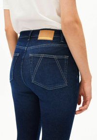 Dunkelblaue Jeans aus Denim mit hohem Bund, kontrastierenden gelben Nähten, einem Lederetikett mit der Aufschrift "ARMEDANGELS" und schrägen Gesäßtaschen.