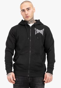 Sort zip-up hoodie med en frontlomme, der har et fremtrædende hvidt "TAPOUT" logo. Fremstillet af blødt stof med en glat overflade.