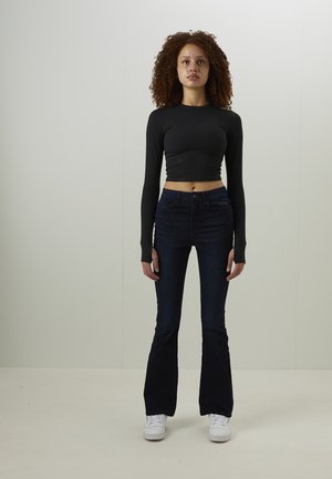 Svart långärmad croppad topp tillsammans med mörkblå utsvängda jeans och vita sneakers. Modellen står frontalt mot en ljus bakgrund.