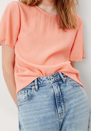 Femme portant un haut corail clair à manches courtes avec des bords festonnés, rentré dans un jean en denim bleu clair taille haute.