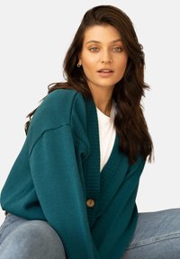 Akwamarynowy sweter z dzianiny charakteryzuje się głębokim dekoltem w kształcie V, ribbowanym wzorem oraz zapięciem na drewniane guziki, w połączeniu z białym T-shirtem i niebieskimi dżinsami.