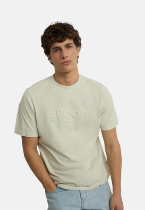 Joven con cabello castaño rizado que lleva una camiseta beige clara con relieve y vaqueros azul claro, de pie con una mano en el bolsillo.
