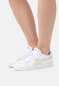 Zapatillas blancas con una parte superior de cuero texturizado, que presentan una franja lateral de un beige pálido y acentos en rosa en la lengüeta. Cintas planas y suela de goma.