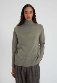 Donna con capelli lunghi e ricci che indossa un maglione a collo alto verde oliva e pantaloni a gamba larga a quadri scuri, su sfondo bianco.