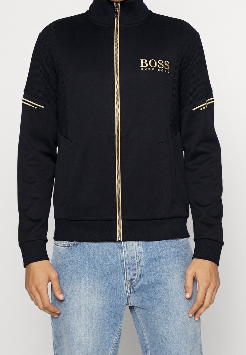 Veste noire zippée avec une fermeture éclair dorée et le logo "BOSS HUGO BOSS" sur la poitrine, portée avec un jean bleu clair.