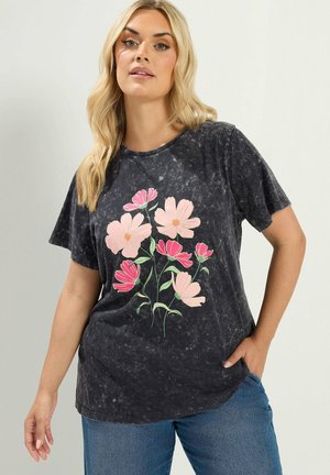 Frau mit langen blonden Haaren, die ein schwarz marmoriertes T-Shirt mit pinken und roten Blumenmustern und blaue Jeans trägt, posiert mit einer Hand in der Tasche.