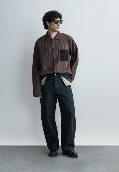 Άνδρας με σγουρά μαλλιά που φοράει μαύρα γυαλιά ηλίου, καφέ ριγέ πουκάμισο με υφασμένη τσέπη, μαύρο φαρδύ παντελόνι με καρό λεπτομέρειες και μαύρα παπούτσια.