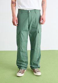 Pantalons cargo verts en tissu texturé, avec des poches latérales, une fermeture à bouton et une coupe décontractée. Portés avec des baskets marron.