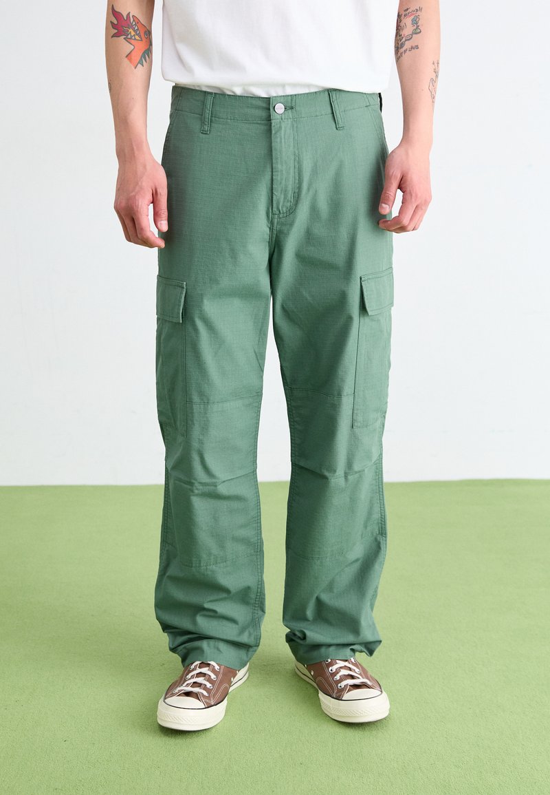 Pantalons cargo verts en tissu texturé, avec des poches latérales, une fermeture à bouton et une coupe décontractée. Portés avec des baskets marron.