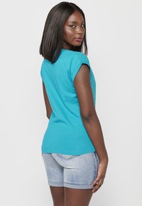 Türkiz kétrészes ujjú felső, részletes varrással a vállakon, világos farmer sorttal párosítva. Zökkenőmentes anyag és testhezálló kialakítás.