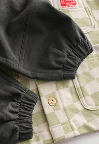 Detail zeleno-beige kockovanej košele s elastickými tmavošedými rukávmi a dreveným gombíkom. Textúrovaná bavlnená látka.