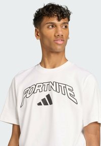 White cotton t-shirt featuring a large black "FORTNITE" logo and Adidas three-stripe design. Short sleeves and round neckline.