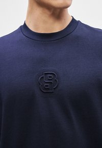 Ναυτικό φούτερ με στρογγυλή λαιμόκοψη, υφασμάτινη υφή και ανάγλυφο λογό τυπο στο στήθος, χαρακτηρίζεται από έναν απλό και καθαρό σχεδιασμό.