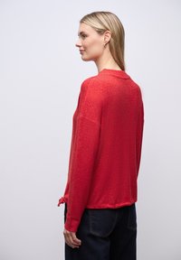 Czerwony sweter dziany o fakturowanym wykończeniu, z wysokim dekoltem i związanym akcentem z tyłu, w połączeniu z ciemnoniebieskimi dżinsami.