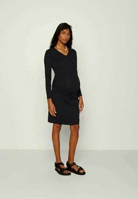 Mulher grávida vestindo um vestido preto justo de manga longa e sandálias pretas, de pé contra um fundo claro e simples.