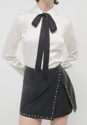 Witte satijnen blouse met lange mouwen en zwarte stropdas, gecombineerd met een zwarte asymmetrische minirok versierd met zilveren studs langs de randen.