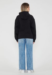 Felpa nera con cappuccio dal taglio rilassato, abbinata a jeans a gamba larga blu chiaro con orli sfrangiati e sneakers bianche.