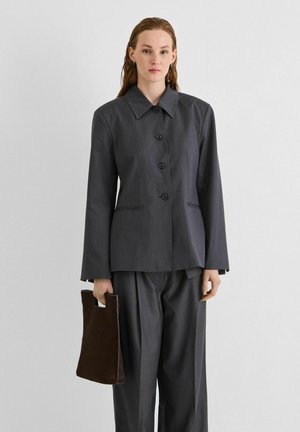Une femme présente une veste anthracite boutonnée sur le devant et un pantalon à jambes larges. Elle porte un cabas brun foncé sur un fond clair et uni.