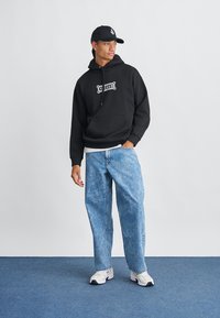 Zwarte hoodie met witte graphic, lichtblauwe versleten denim jeans en witte sneakers met zwarte accenten. Man staat tegen een effen achtergrond.
