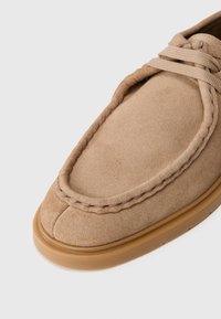 Smėlio spalvos zomšiniai mocasinai su apvalia nosimi, dekoratyvinėmis siūlėmis aplink kraštą ir lanksčiu guminiu padu.