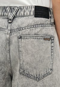 Πίσω όψη τζιν παντελονιού από ανοιχτό γκρι acid-wash denim, με δεξιά πίσω τσέπη που φέρει μια μικρή ετικέτα της μάρκας Volcom και μια μαύρη επιφάνεια πάνω από τη μέση.