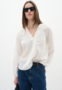 Blusa bianca a maniche lunghe con scollo a V, caratterizzata da sottili strisce verticali. Abbinata a jeans blu scuro e accessoriata con occhiali da sole neri.