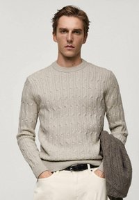 Helle beige Kabelstrickpullover mit Rundhalsausschnitt und gerippten Bündchen, kombiniert mit cremefarbenen Hosen. Graue strukturierte Jacke lässig über dem Arm getragen.