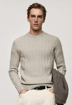 Helle beige Kabelstrickpullover mit Rundhalsausschnitt und gerippten Bündchen, kombiniert mit cremefarbenen Hosen. Graue strukturierte Jacke lässig über dem Arm getragen.