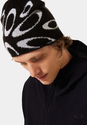 Beanie en tricot noir avec des motifs circulaires blancs. S'ajuste parfaitement à la tête. Petite étiquette de marque sur le côté. Porté avec une veste zippée noire.