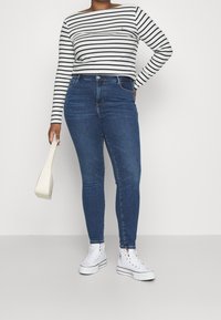 Pruhovaný dlouhý rukáv v šedé a námořnické barvě, módní modré džíny s vysokým pasem a bílé vysoké tenisky. Drží bílou kabelku.