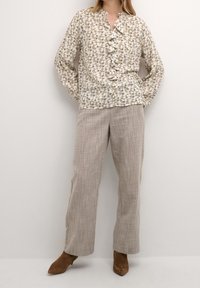 Biała bluzka w kwiaty z falbaniastym przodem, długimi rękawami i okrągłym dekoltem, zestawiona z beżowymi, teksturowanymi spodniami typu wide-leg i brązowymi botkami na kostkę.