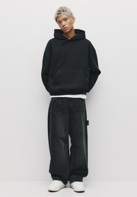 Hanorac oversized negru cu buzunar frontal, asociat cu blugi largi de culoare gri închis, cu accente distruse. Adidași albi completează look-ul.