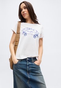 Νεαρή γυναίκα που φοράει λευκό μπλουζάκι με σχέδιο, τζιν φούστα και ταμπά τσάντα ώμου, στέκεται με τα χέρια στις τσέπες μπροστά σε απλό φόντο.