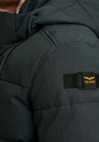 Veste matelassée vert foncé avec un design cousu, col en polaire et un patch noir avec le logo "PME Legend" sur l'épaule.