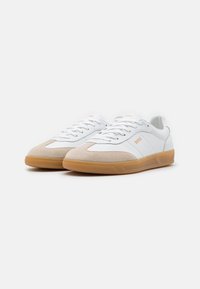 White sneakers made of leather and suede, featuring a beige toe cap, orange branding, and a gum rubber sole. Classic low-top design.