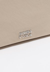 Beżowa torba z materiału o gładkiej fakturze i tłoczonym logo "BIMBA Y LOLA" w kolorze srebrnym w prawym dolnym rogu.