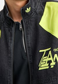 Närbild av en person som bär en svart och neon gul Adidas Team-jacka med en frontdragkedja som är delvis öppen.
