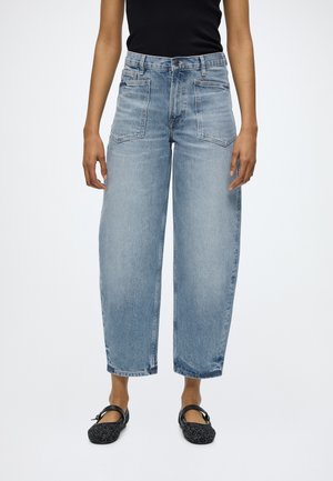 Person trägt hellblaue, hoch taillierte, weit geschnittene Jeans mit großen Fronttaschen, ein schwarzes ärmelloses Oberteil und schwarze Mary Jane Ballerinas.