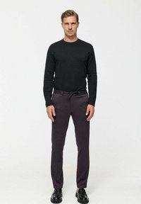 Man standing, wearing a black long-sleeve shirt, dark purple drawstring pants, and black shoes against a plain light background.