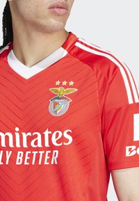 Červený športový dres s výstrihom do V, s textúrovanými pruhmi, klubovým emblémom s orlom a bielymi akcentmi na golieri a rukávoch.