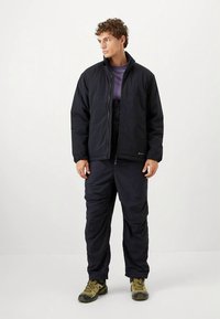 Black insulated jacket with high collar, zip front, paired with matching dark pants and yellow hiking boots. Fabric appears durable and textured.