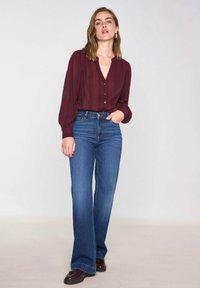 Blouse bordeaux avec un décolleté en V et des boutons, associée à un jean bleu taille haute et évasé. Le modèle porte des chaussures foncées, se tenant devant un fond neutre.