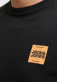 Schwarzes Baumwoll-T-Shirt mit einem quadratischen orangen Etikett, auf dem "Jack & Jones" in schwarzer Schrift steht; glatte Textur und figurbetonter Schnitt.